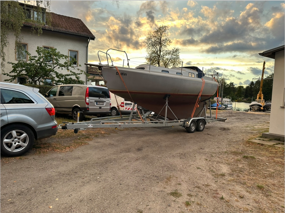 Ein Auto zieht ein Boot auf einem Anhänger entlang einer Straße, mit Bäumen und einem klaren Himmel im Hintergrund.